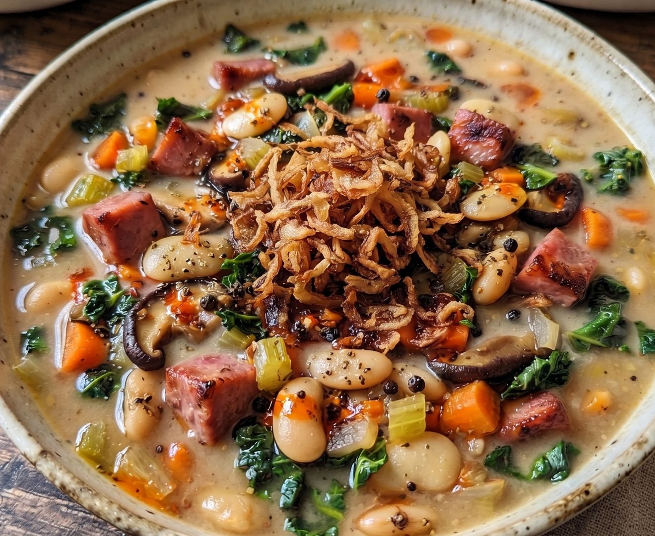 Bowl of ham and bean soup with white beans, diced ham, carrots, celery, mushrooms, and kale, topped with fried shallots