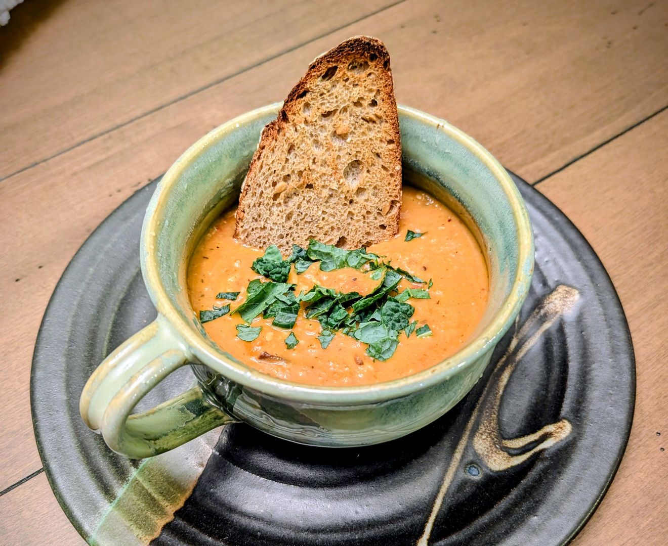 Roasted tomato soup topped with chopped kale in a green ceramic mug, with a slice of toasted bread tucked in the side